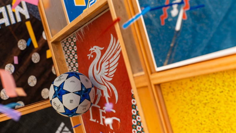 Blue-and-white football floating beside a red Liverpool panel with colourful confetti and gold frames in the One Two artwork.