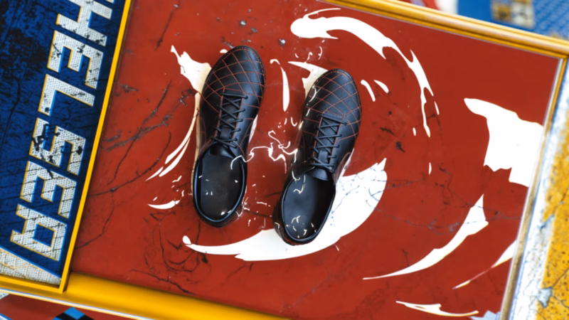 Pair of black football boots on a red panel with white brush strokes beside Chelsea lettering in the One Two graphics.